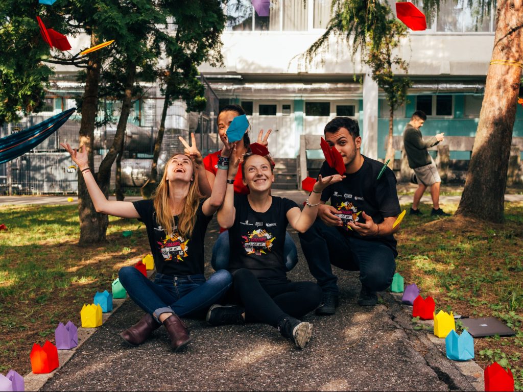 Volunteer students sitting on park path, smiling and throwing colourful origami in the air.