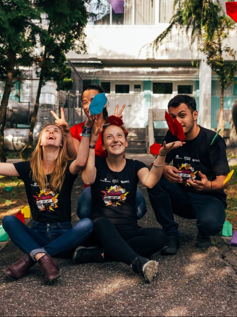 Volunteer students sitting on park path, smiling and throwing colourful origami in the air.