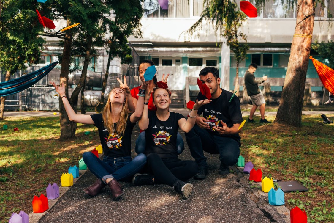 Volunteer students sitting on park path, smiling and throwing colourful origami in the air.